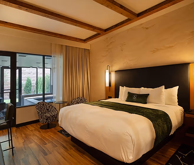 Elegant king-size bed with a green throw in a modern guest room at The Swiftwater hotel, featuring a view of the private indoor plunge pool through large windows.