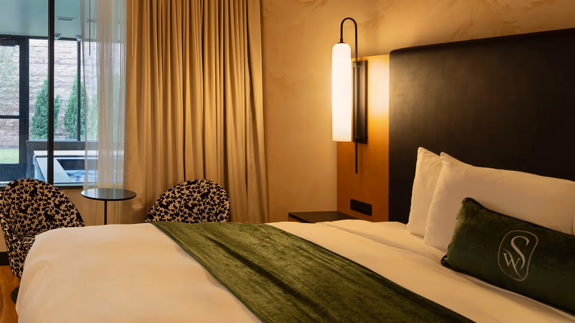 Close-up of a guest room at The Swiftwater hotel showing a cozy bed with plush white pillows, a green accent cushion, and contemporary wall lighting.