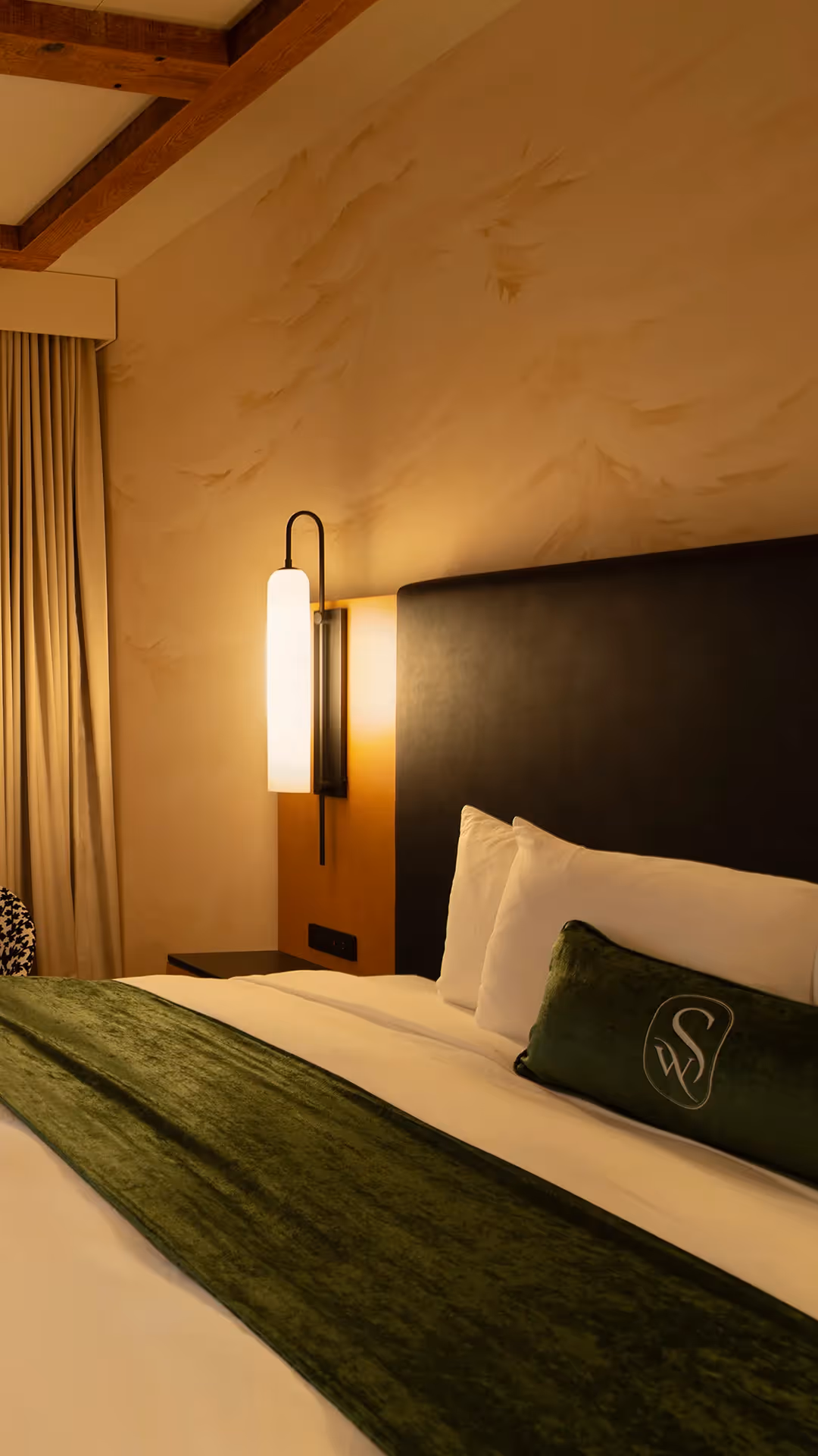 Close-up of a guest room at The Swiftwater hotel showing a cozy bed with plush white pillows, a green accent cushion, and contemporary wall lighting.