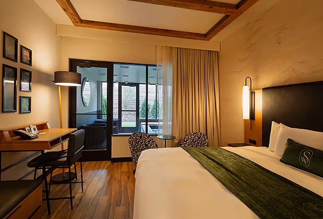 Spacious hotel room at The Swiftwater featuring a wooden work desk, designer armchair, and warm lighting next to floor-to-ceiling windows.
