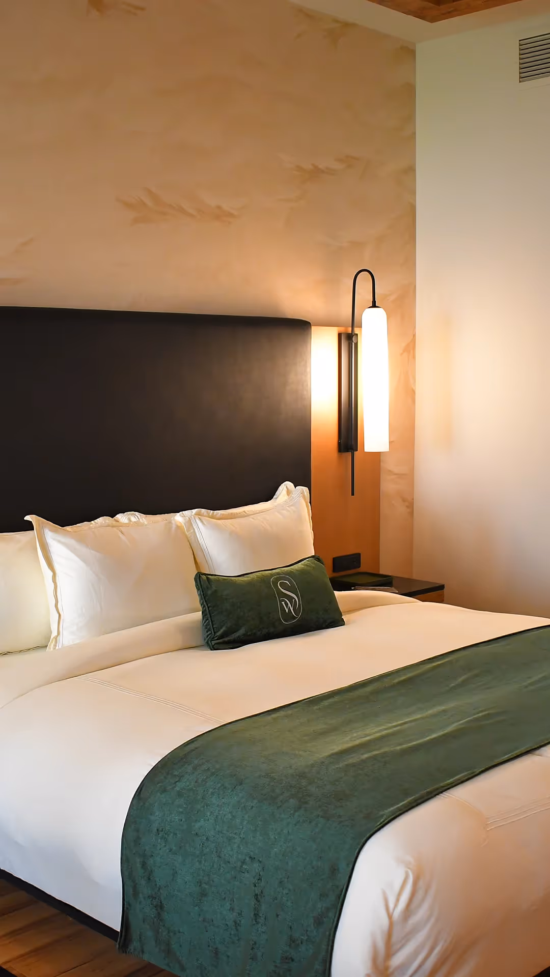 Close-up of a luxurious bed at The Swiftwater with crisp white linens, a dark headboard, and a signature green velvet lumbar pillow against a decorative wall.