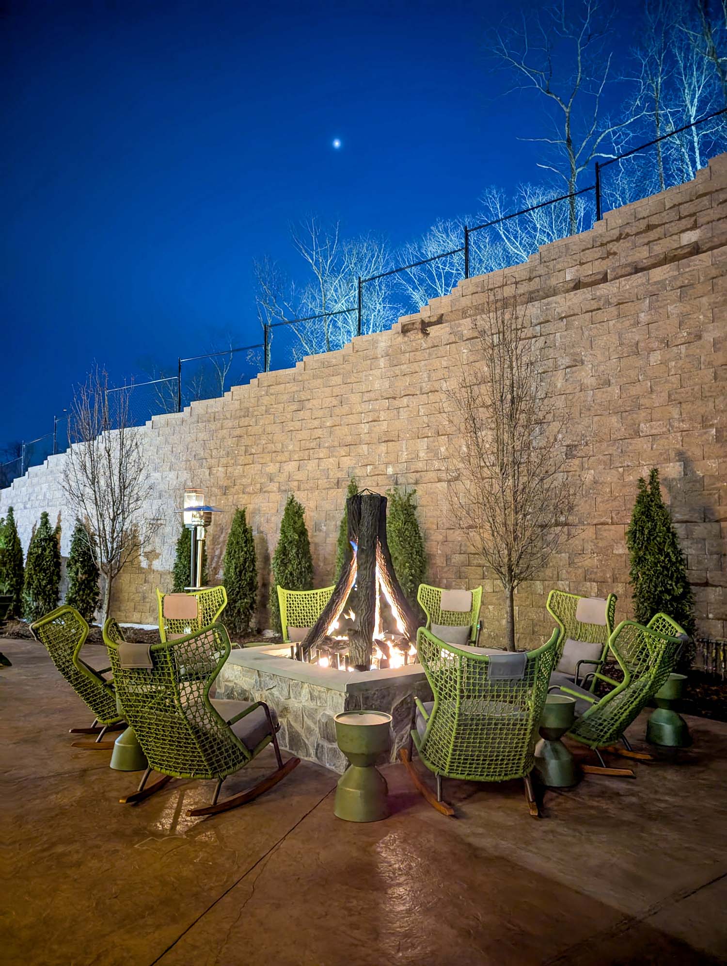 A large stone outdoor fire pit at The Swiftwater Hotel featuring tipi-style sculpted logs and warm flames, surrounded by modern lounge chairs under soft evening lighting.