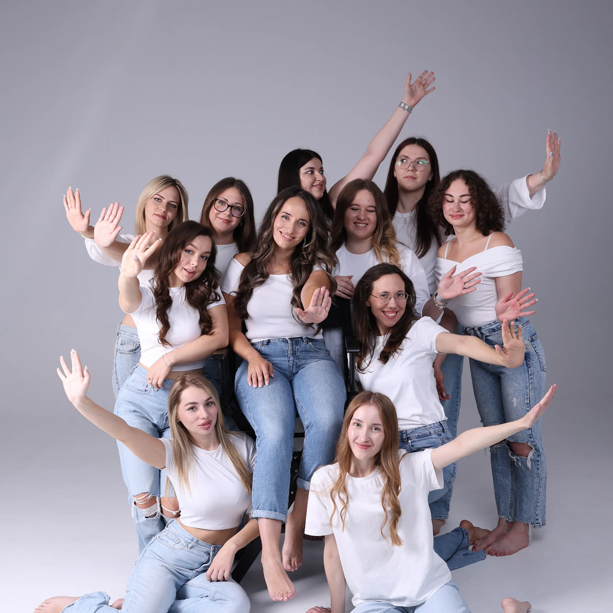 Grupa jedenastu młodych kobiet ubranych w białe koszulki i niebieskie dżinsy, pozujących z uśmiechami i uniesionymi rękami na szarym tle.