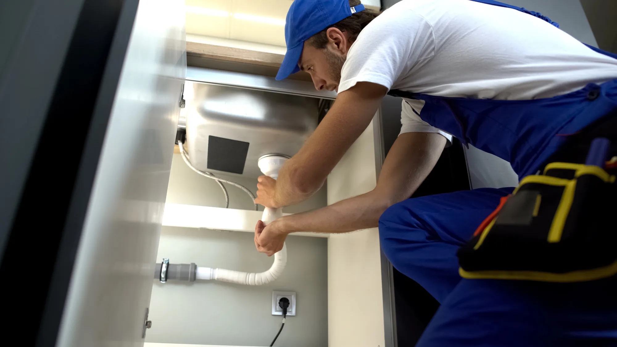 Plumber in blue overalls and cap installing or fixing a white pipe under a sink.