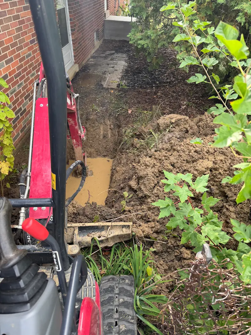 Excavator digging a muddy trench next to a brick house with green shrubs nearby.