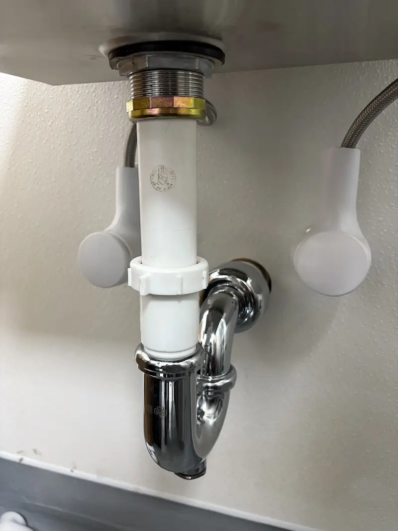 White and chrome P-trap plumbing pipe assembly under a sink against a white textured wall.