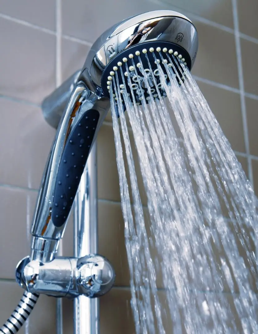 Close-up of a chrome handheld showerhead with water flowing, mounted on a tiled bathroom wall.