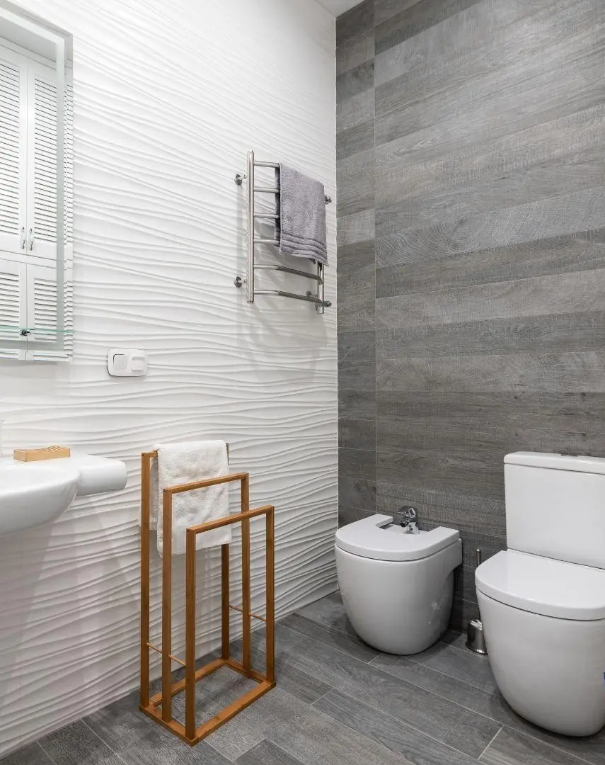 Modern bathroom with white toilet, bidet, textured white wall, gray tiled wall and floor, towel rack with gray towel, and wooden towel stand with white towel.