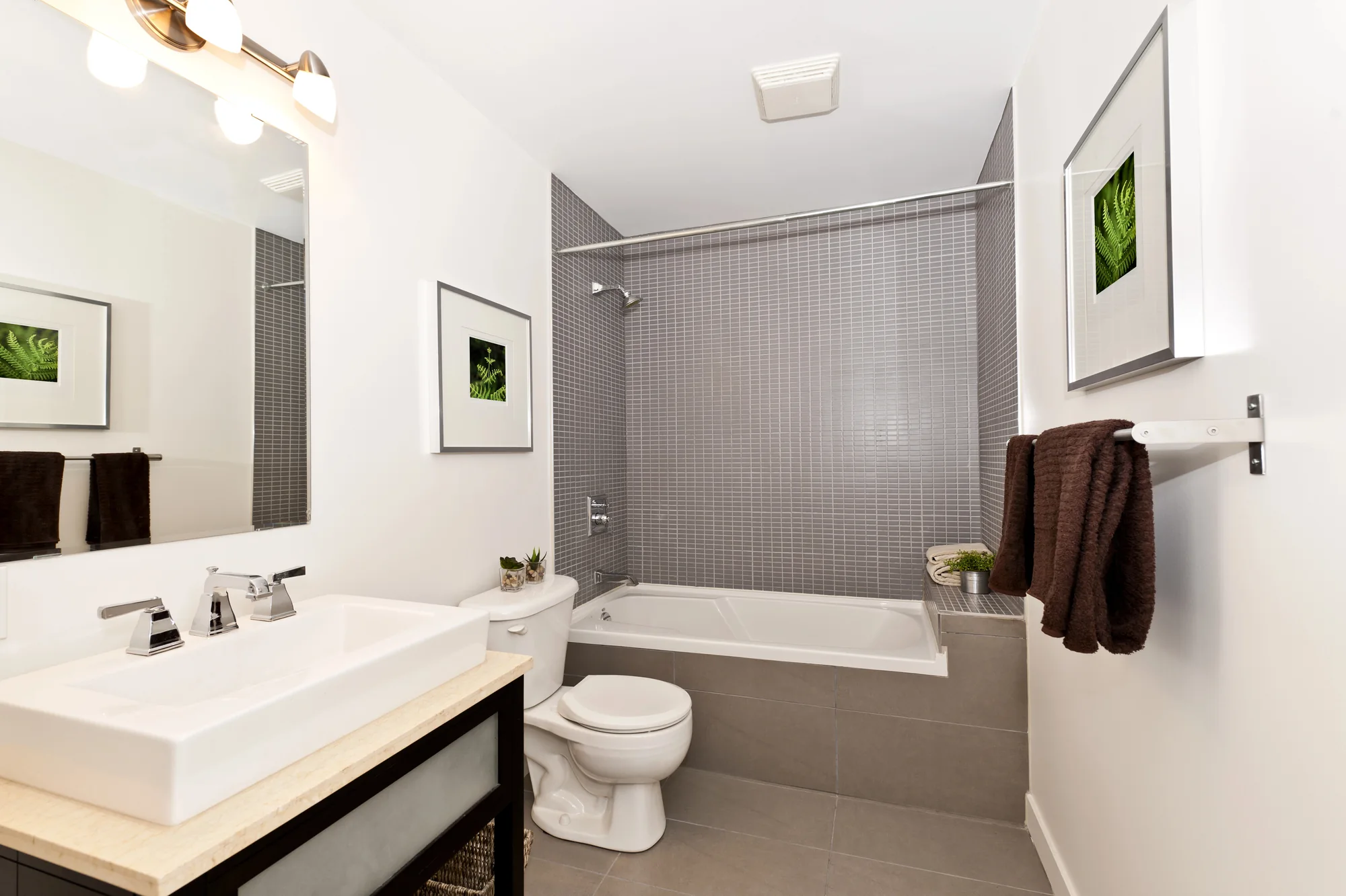 Modern bathroom with white sink, toilet, bathtub with gray tiled wall, brown towels, and framed leaf artwork.