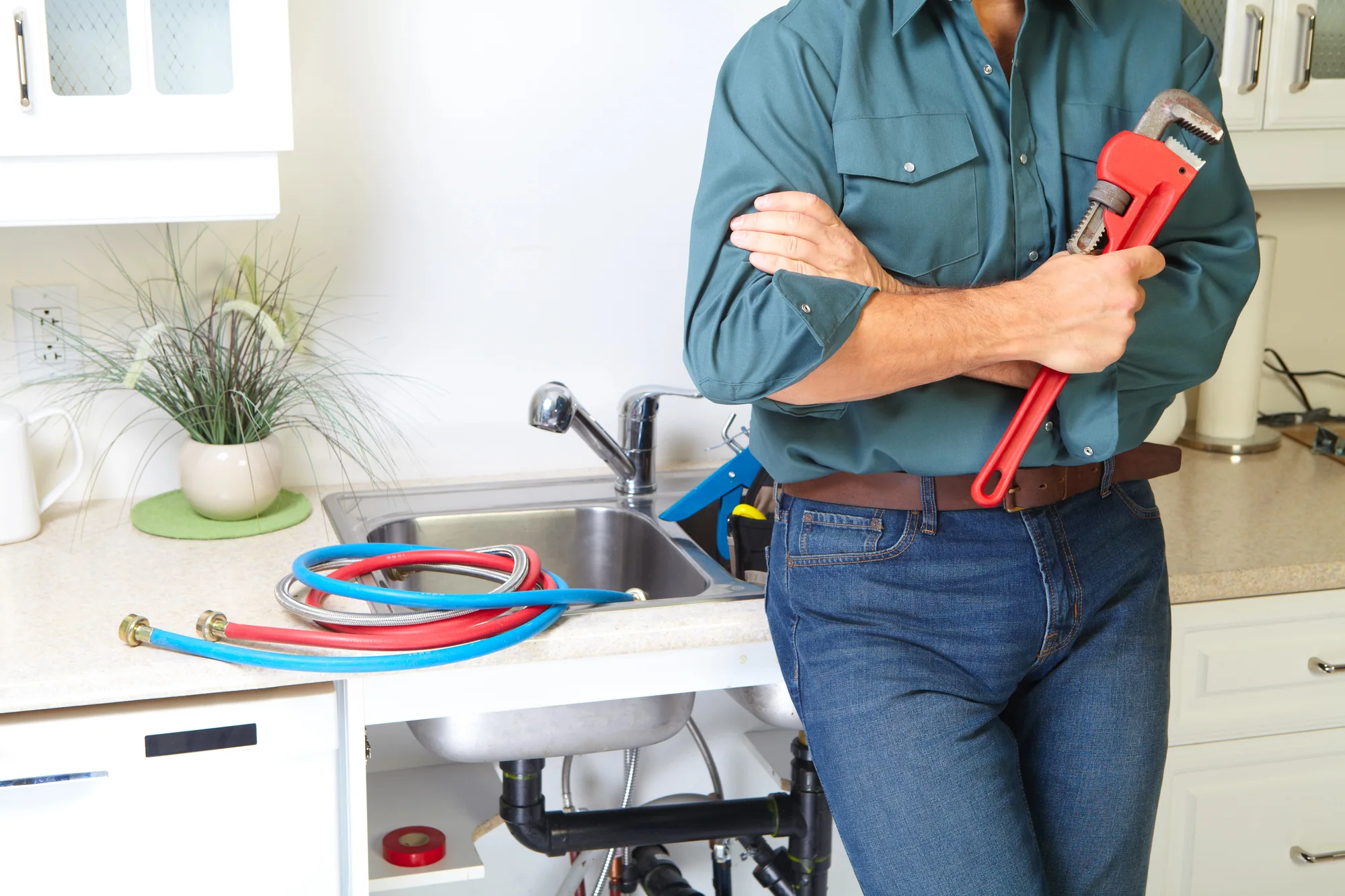 Plumber standing with arms crossed holding a red pipe wrench in a kitchen with an open cabinet under the sink and plumbing hoses on the counter.