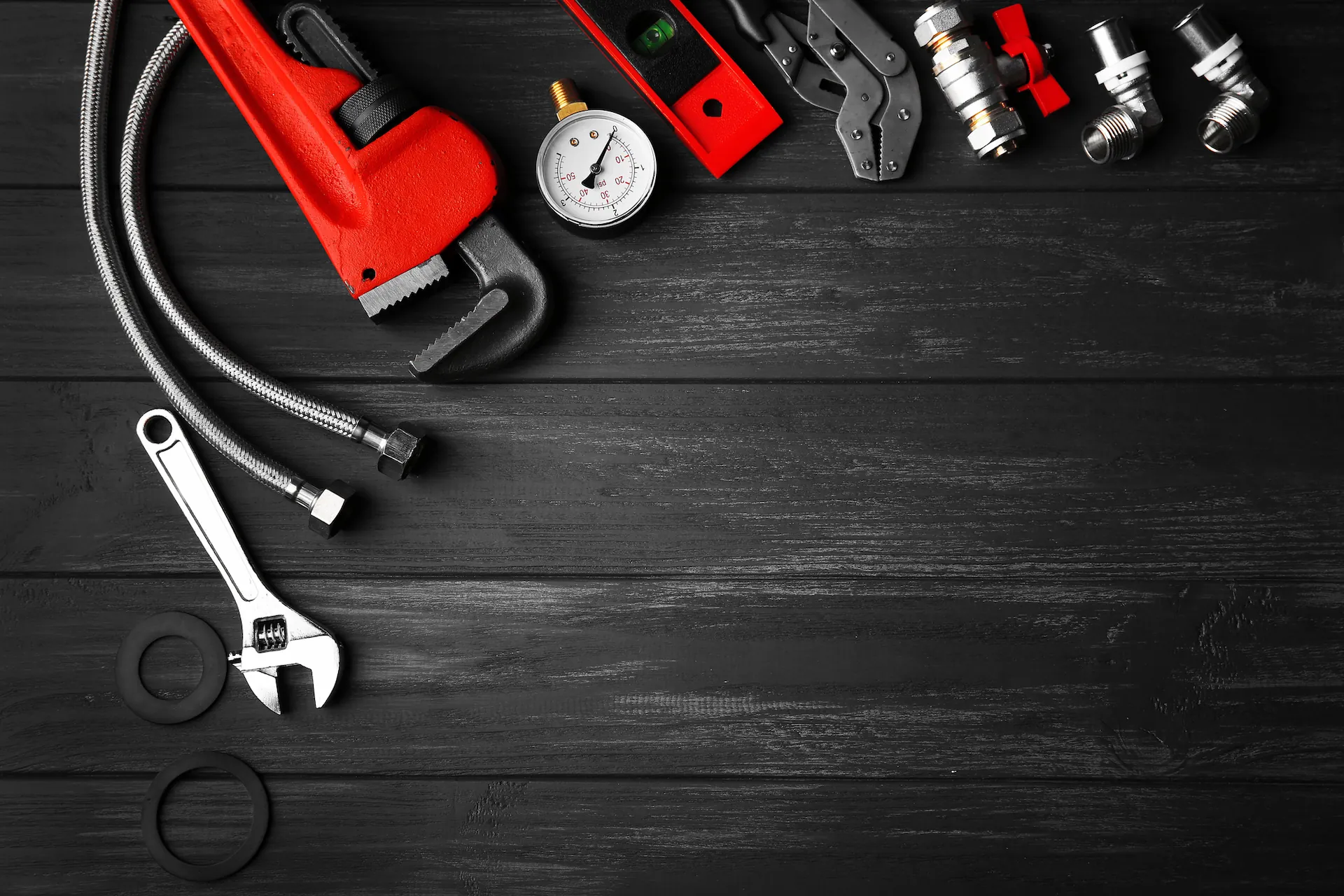 Various plumbing tools and fittings including a red pipe wrench, adjustable wrench, pressure gauge, flexible hoses, rubber washers, and pipe connectors on a dark wooden surface.