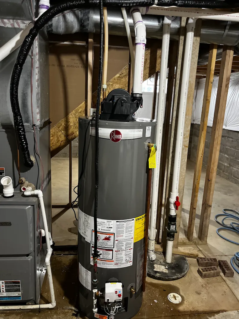 Gray Rheem water heater installed in a basement with PVC pipes and wooden beams around.