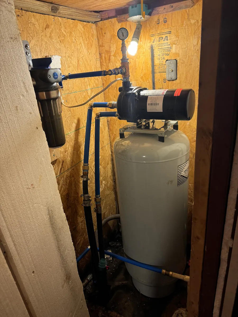 Water pressure tank and pump system installed in a wooden utility closet with exposed pipes and a pressure gauge.
