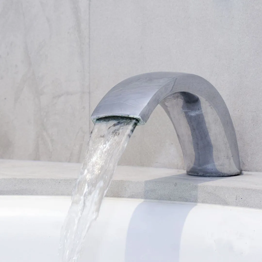 Modern silver faucet with water flowing into a white basin against a gray tiled wall.