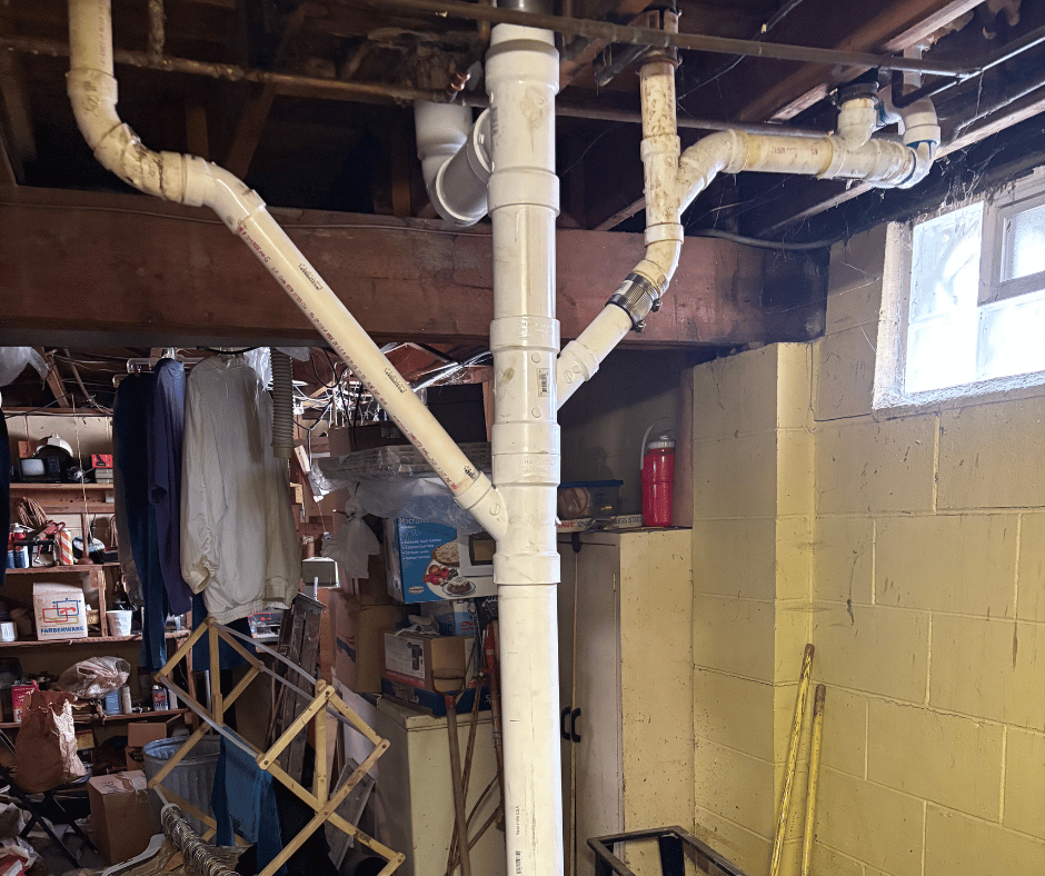 Basement corner with white PVC plumbing pipes, exposed ceiling beams, a small window, and various stored items including clothes and boxes.