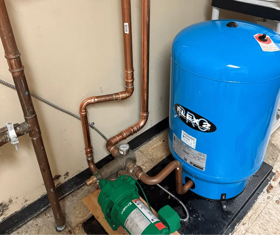 Excavator digging a muddy trench beside a brick house with a pile of dirt and green plants nearby.
