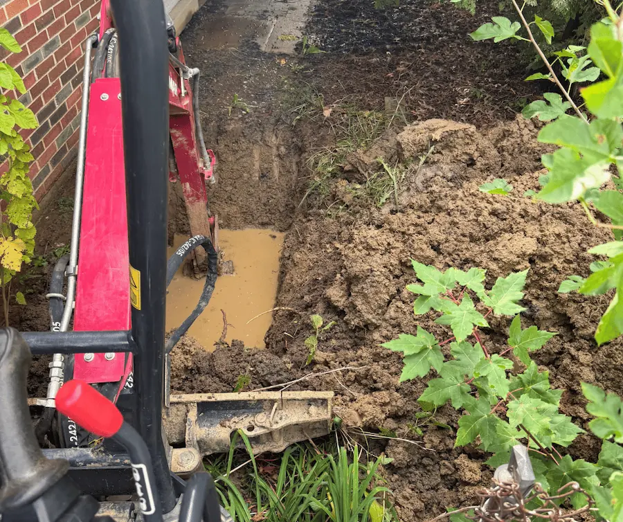 Trench dug in a backyard with a white PVC pipe installed along the trench, surrounded by tools and extra pipe sections on the grass.