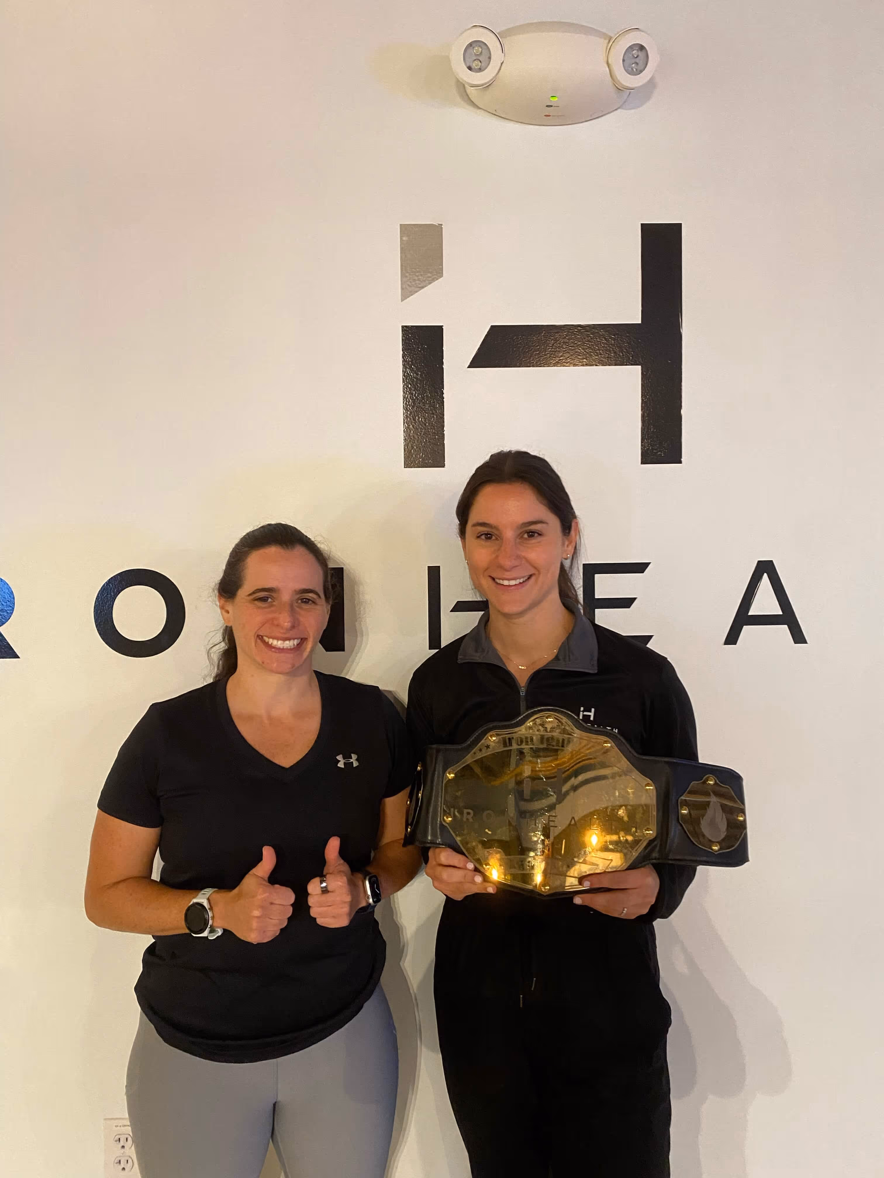 Two smiling women standing in front of a wall with a logo; the woman on the right is holding a large championship belt, and the woman on the left is giving two thumbs up.