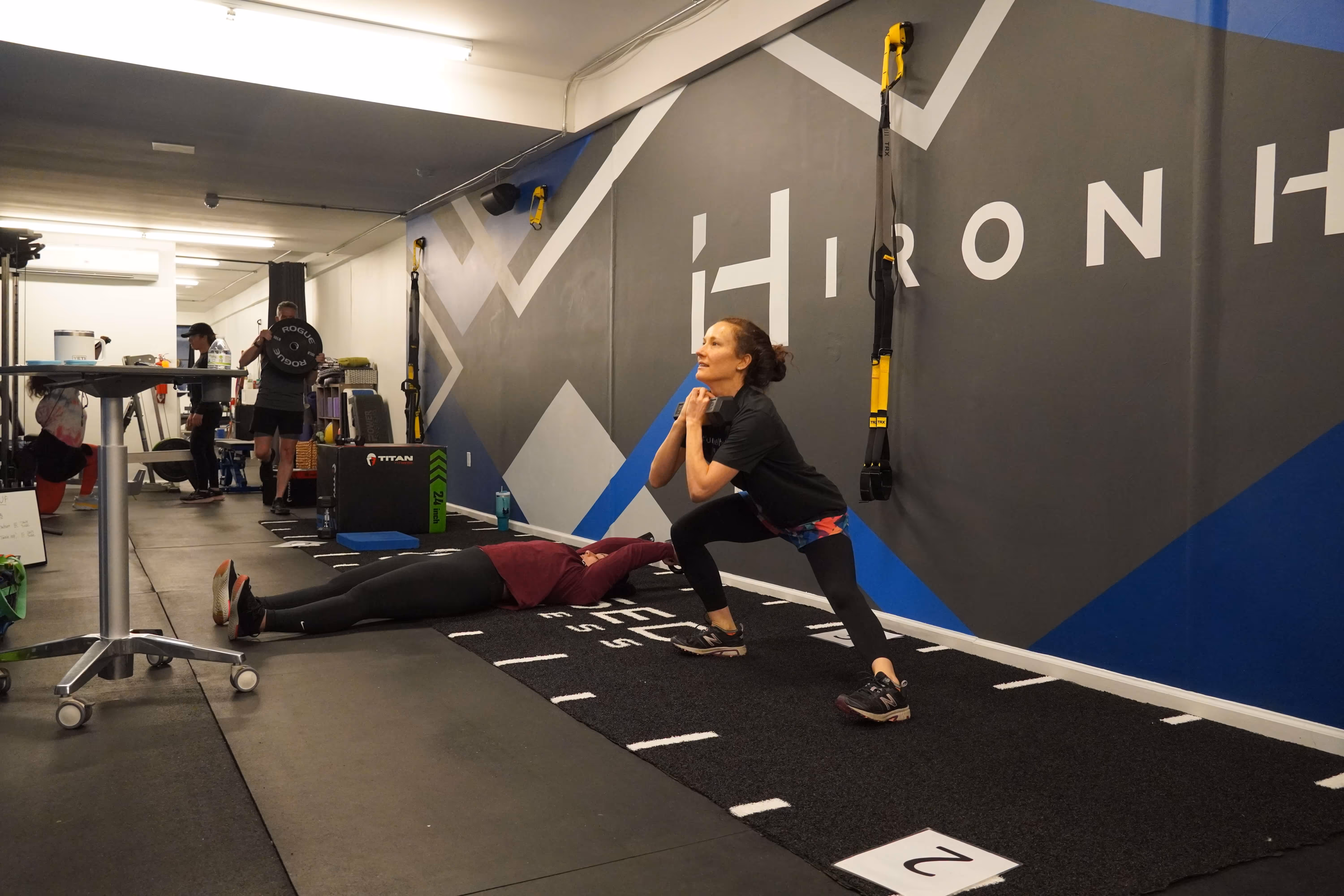 A woman in athletic wear holding a dumbbell and performing a lateral lunge inside a fitness gym.