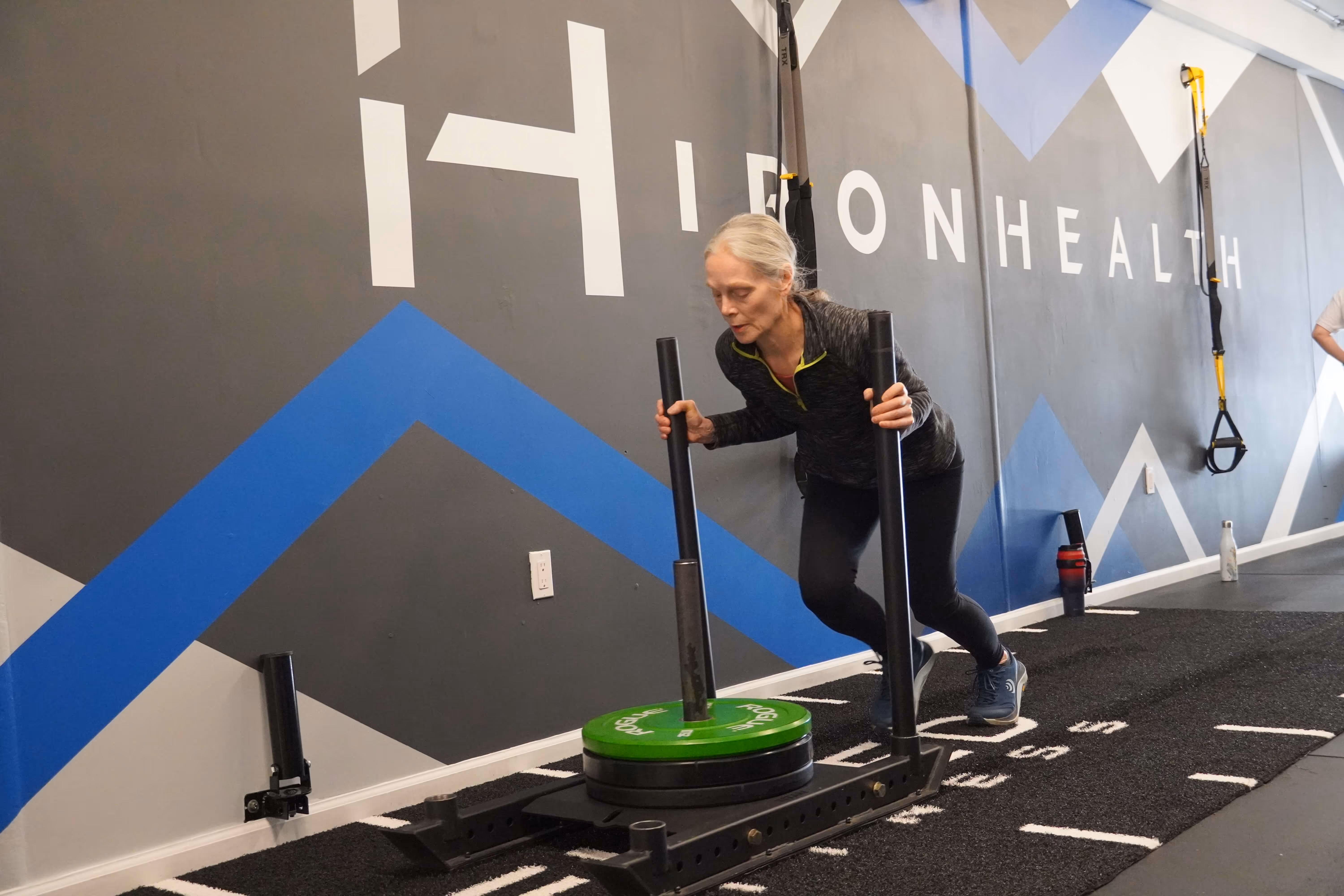 Middle-aged woman pushing a weighted sled on a track inside a gym with a dark wall featuring blue and white geometric designs and the word IONHEALTH.
