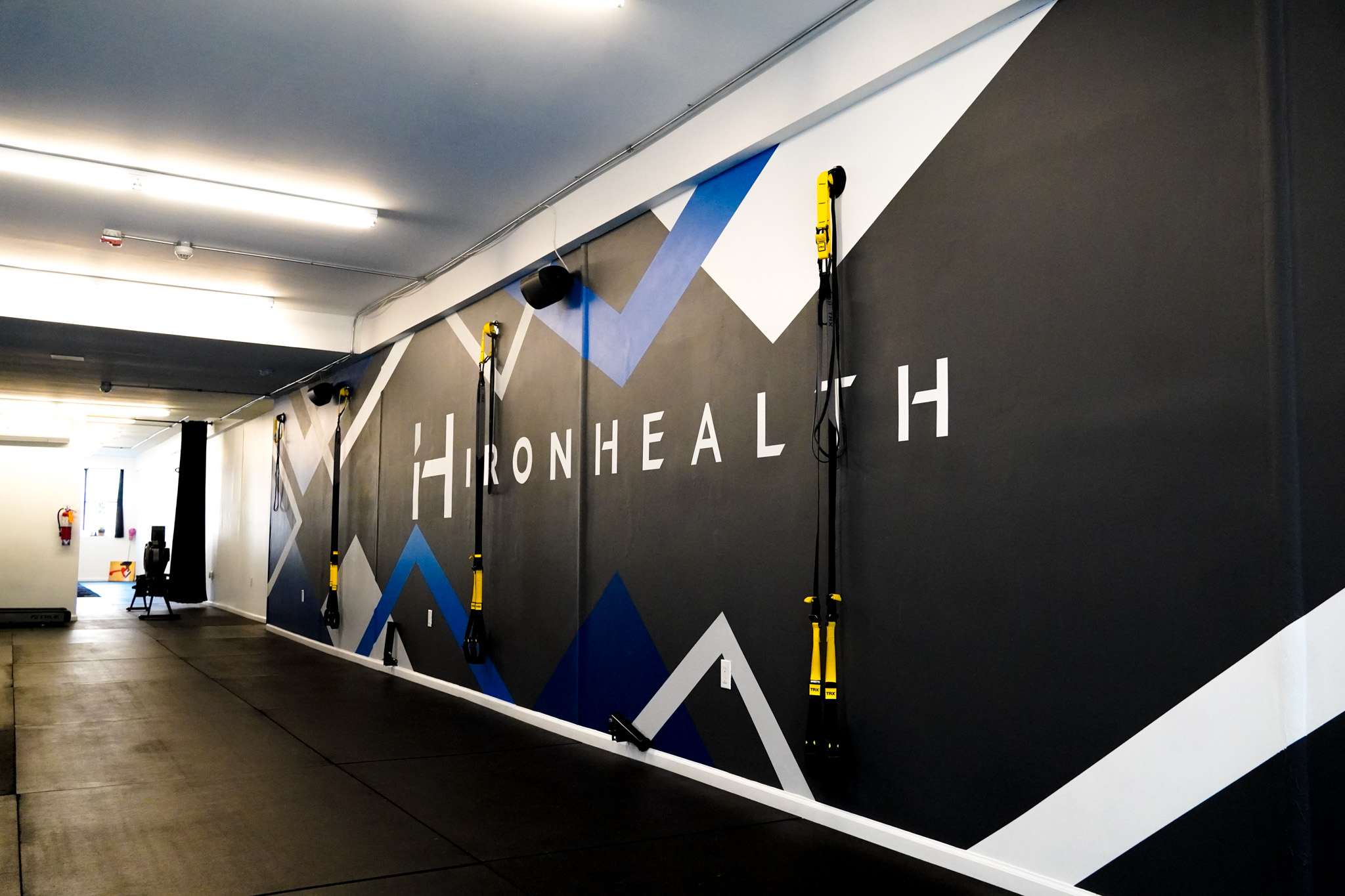 Interior view of a gym with black padded flooring, TRX suspension trainers mounted on a wall with geometric blue, white, and gray patterns, and the text 'IRONHEALTH'.