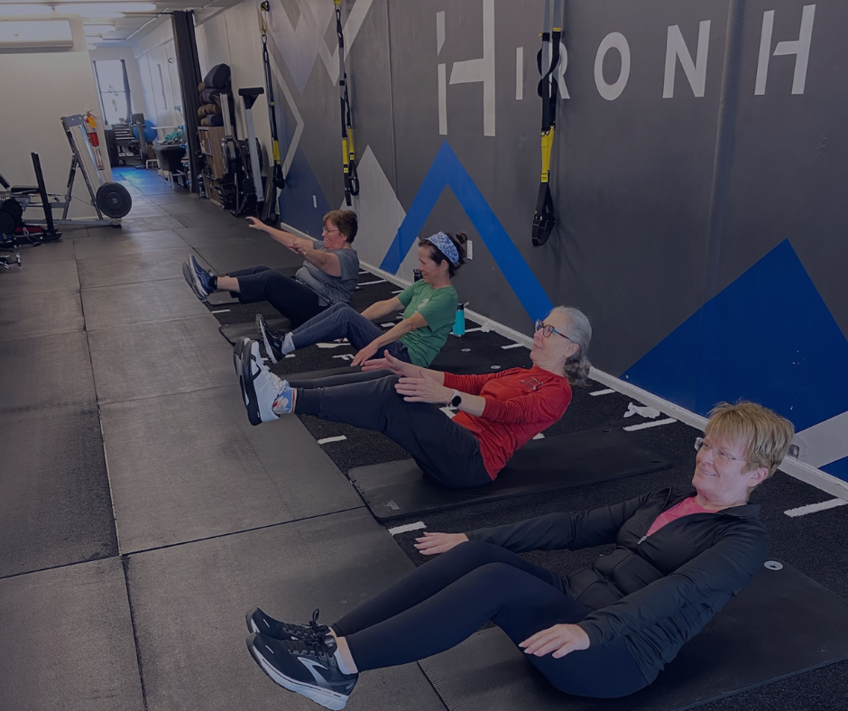 Four women exercising on mats in a gym, performing a seated leg raise workout against a wall with fitness equipment.