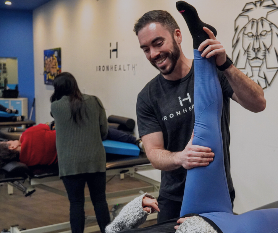 Male physical therapist wearing Iron Health shirt assists a person in blue leggings by stretching their leg upward in a therapy room.