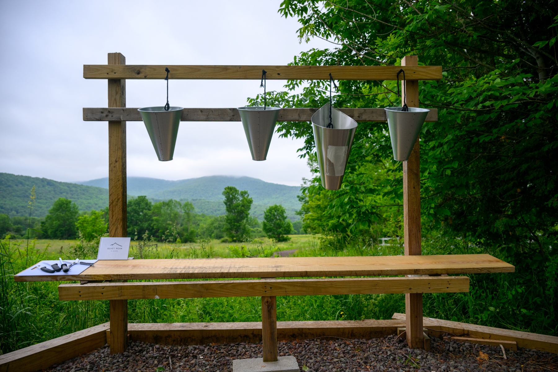 An outdoor farm workstation features a wooden frame with four metal poultry processing cones hanging above a long wooden bench, overlooking a lush green valley and distant mountains.
