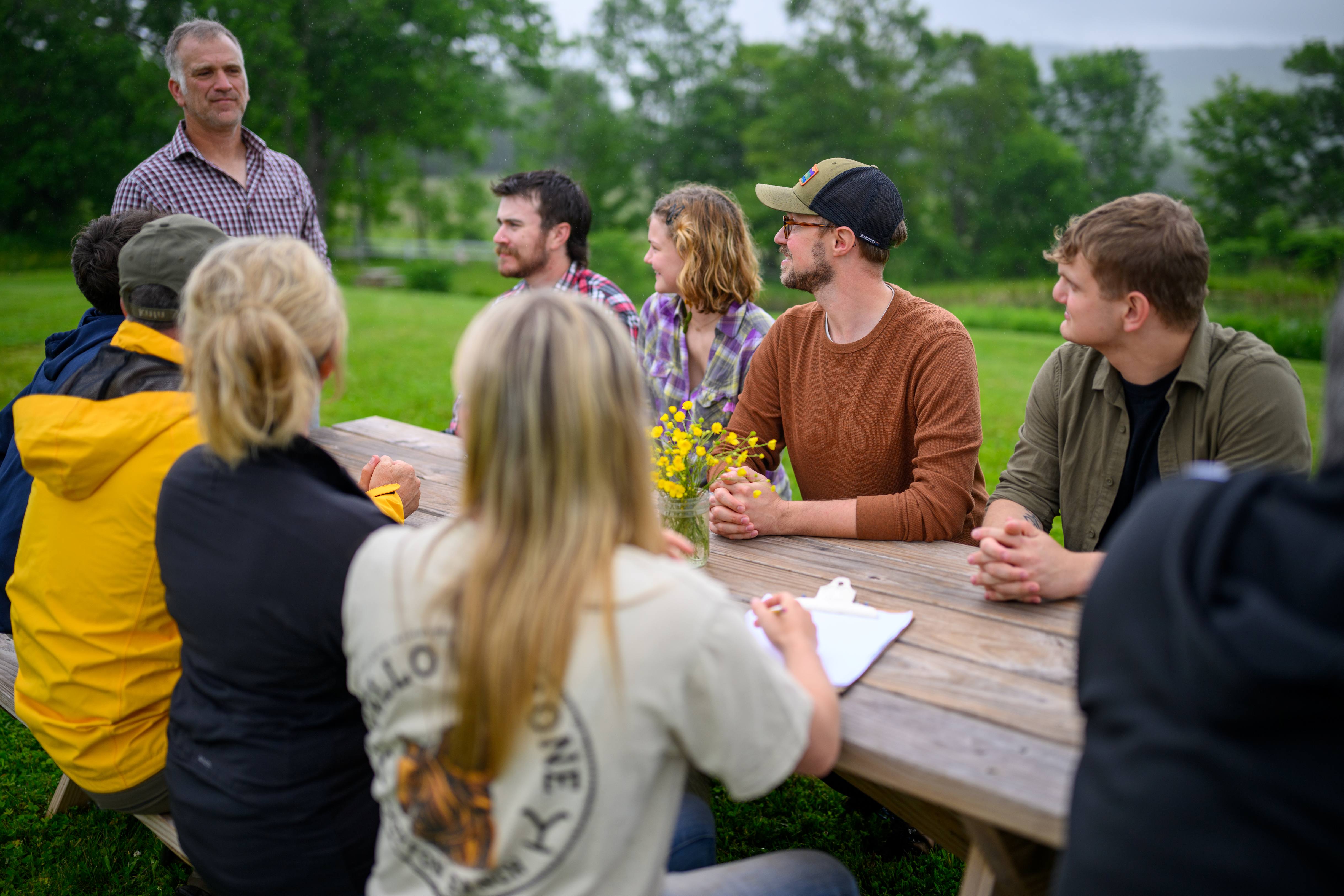A close-up photograph of a white-tailed deer looking directly at the camera with a gentle expression, standing in a grassy meadow with blurred trees in the background under soft light.