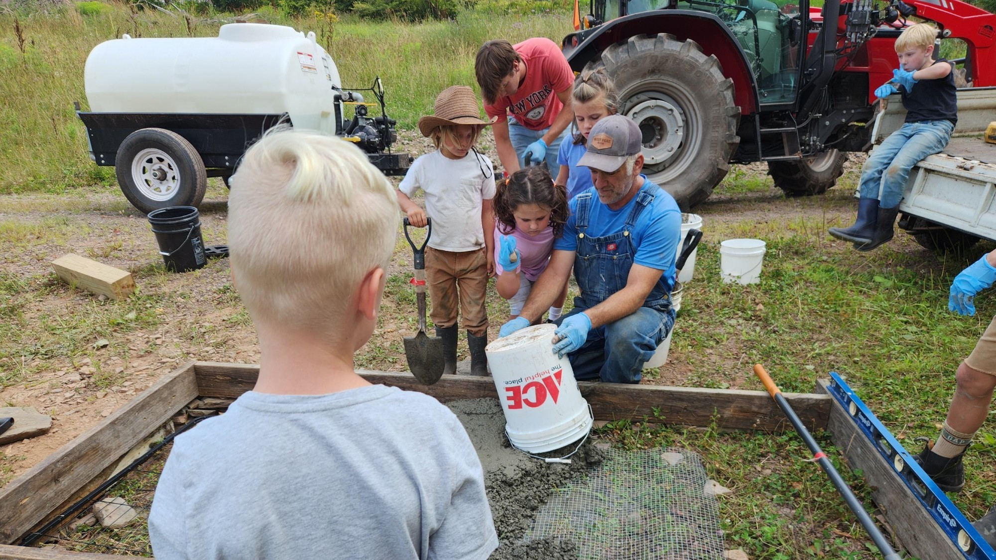 An older man in overalls and a cap pours concrete from a white bucket into a wooden form lined with wire mesh, while several children and another adult watch and assist. In the background are a red tractor and a white water tank on a trailer, all in a grassy field.