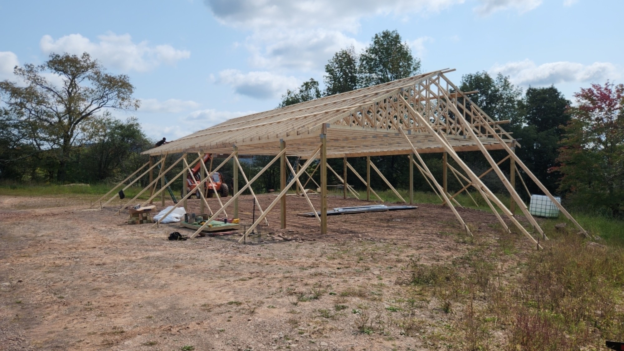 A wide-angle landscape photograph shows the wooden frame of a large pole barn under construction, situated in a cleared dirt field. The frame consists of vertical posts supporting roof trusses. In the background, there are trees and a field under a blue sky with scattered white clouds. A piece of construction equipment is visible on the left side near the frame.