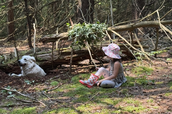 A young girl wearing a floral sun hat and strawberry-patterned boots sits on a mossy patch in a dense forest. Beside her, a large white livestock guardian dog rests in a shallow dirt hole next to a fallen log.