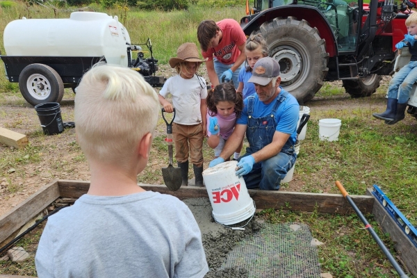 An older man in overalls and a cap pours concrete from a white bucket into a wooden form lined with wire mesh, while several children and another adult watch and assist. In the background are a red tractor and a white water tank on a trailer, all in a grassy field.
