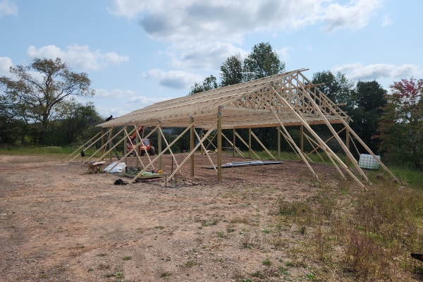 A wide-angle landscape photograph shows the wooden frame of a large pole barn under construction, situated in a cleared dirt field. The frame consists of vertical posts supporting roof trusses. In the background, there are trees and a field under a blue sky with scattered white clouds. A piece of construction equipment is visible on the left side near the frame.