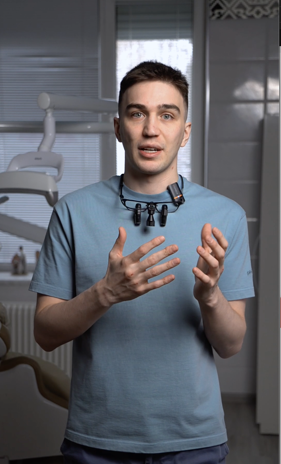 Мужчина в голубой футболке с медицинскими увеличительными очками на шее стоит в стоматологическом кабинете и жестами объясняет что-то.