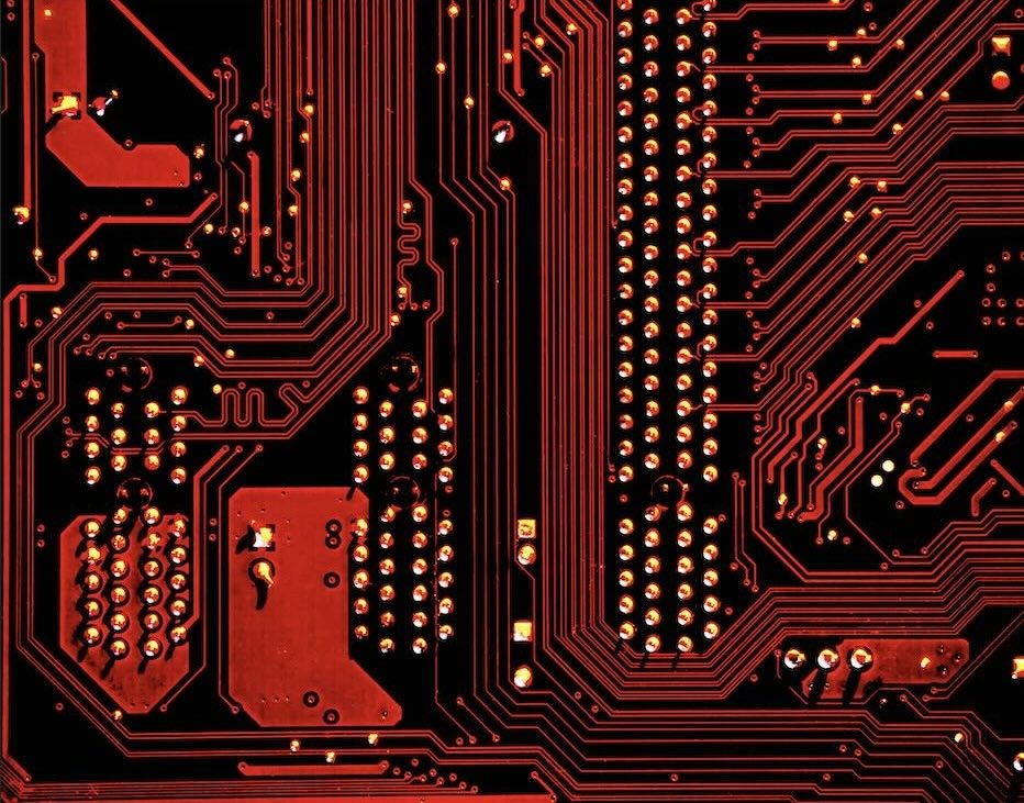 Close-up of a red printed circuit board with multiple solder points and intricate circuitry paths on a black background.