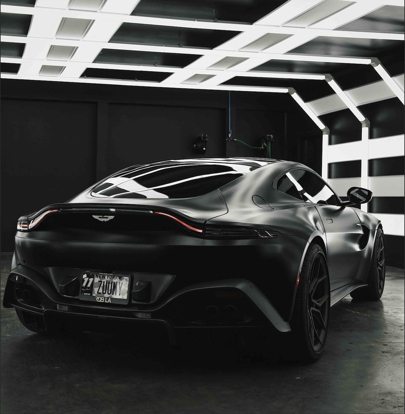 Rear three-quarter view of a black Aston Martin sports car in a modern garage with illuminated ceiling panels.