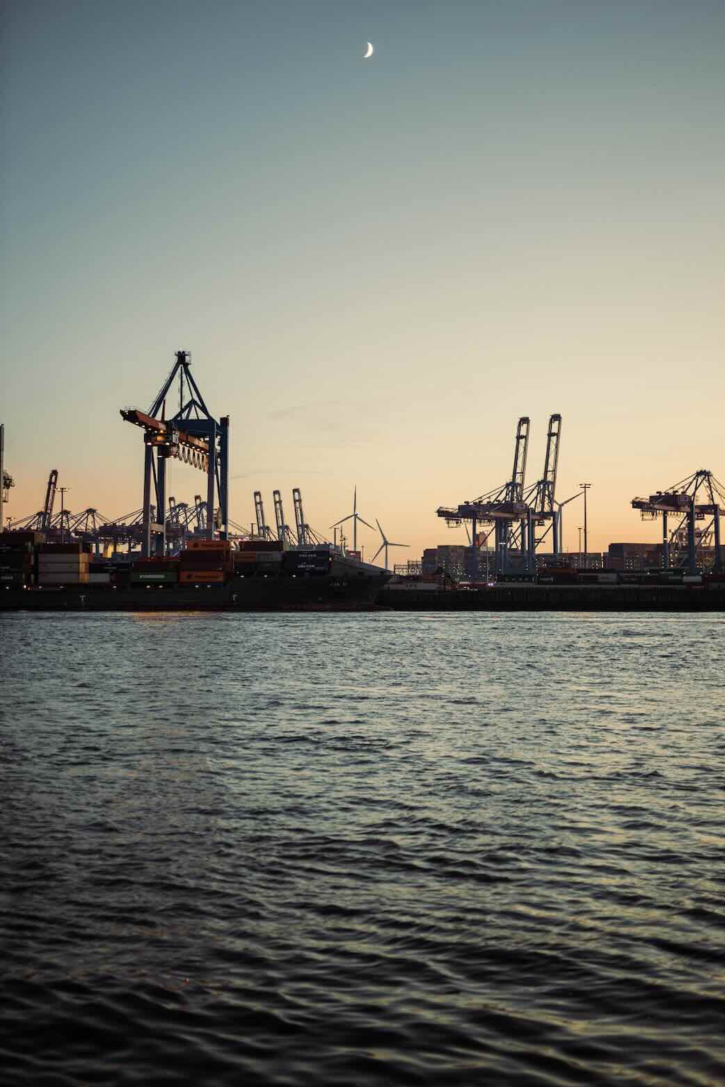 Port at dusk with cranes, shipping containers, wind turbines, and a crescent moon above calm water.