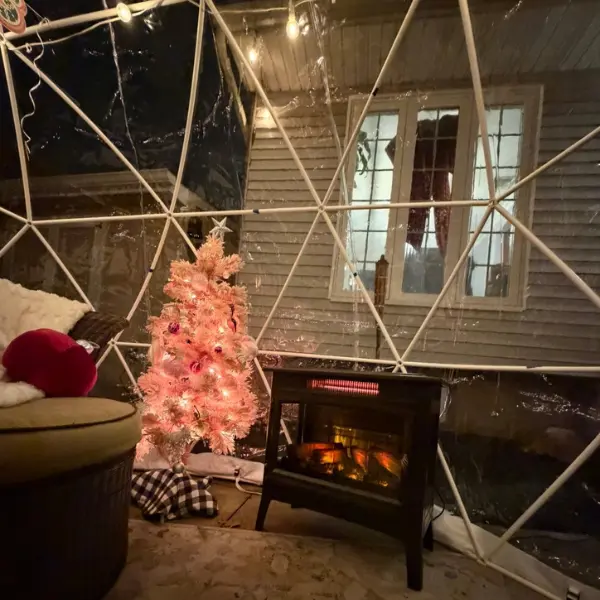 Cozy indoor igloo setup with a small pink-lit Christmas tree, a black electric heater mimicking a fireplace, and a cushioned chair with red and white pillows, all inside a transparent dome structure at night.