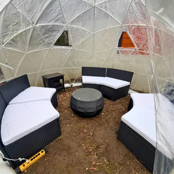 Interior of a transparent geodesic dome with black wicker curved sofas with white cushions arranged around a matching round wicker table on a dirt floor.