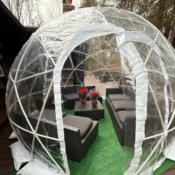 Clear inflatable igloo dome with a zipper door on a wooden deck, containing black wicker patio furniture with gray cushions and two small tables holding red poinsettia plants on artificial grass.