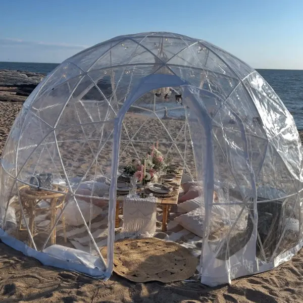 Clear geodesic dome set up on a sandy beach near the ocean, with a low wooden table inside surrounded by cushions and decorated with flowers and candles for a cozy outdoor dining experience.