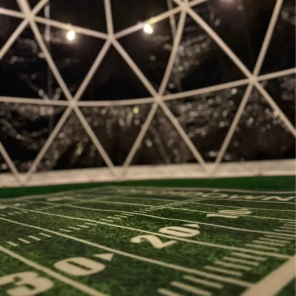 Close-up view of an indoor small football field with artificial green turf and white yard lines inside a dome structure with triangular framing and string lights.