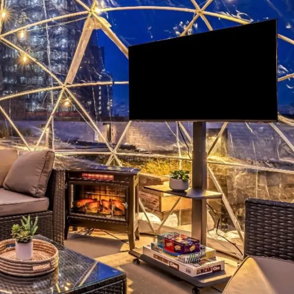 Cozy indoor seating area inside a transparent geodesic dome at night, featuring a TV on a stand, a small electric fireplace heater, board games stacked on a shelf below the TV, rattan furniture, and potted plants, illuminated by string lights.