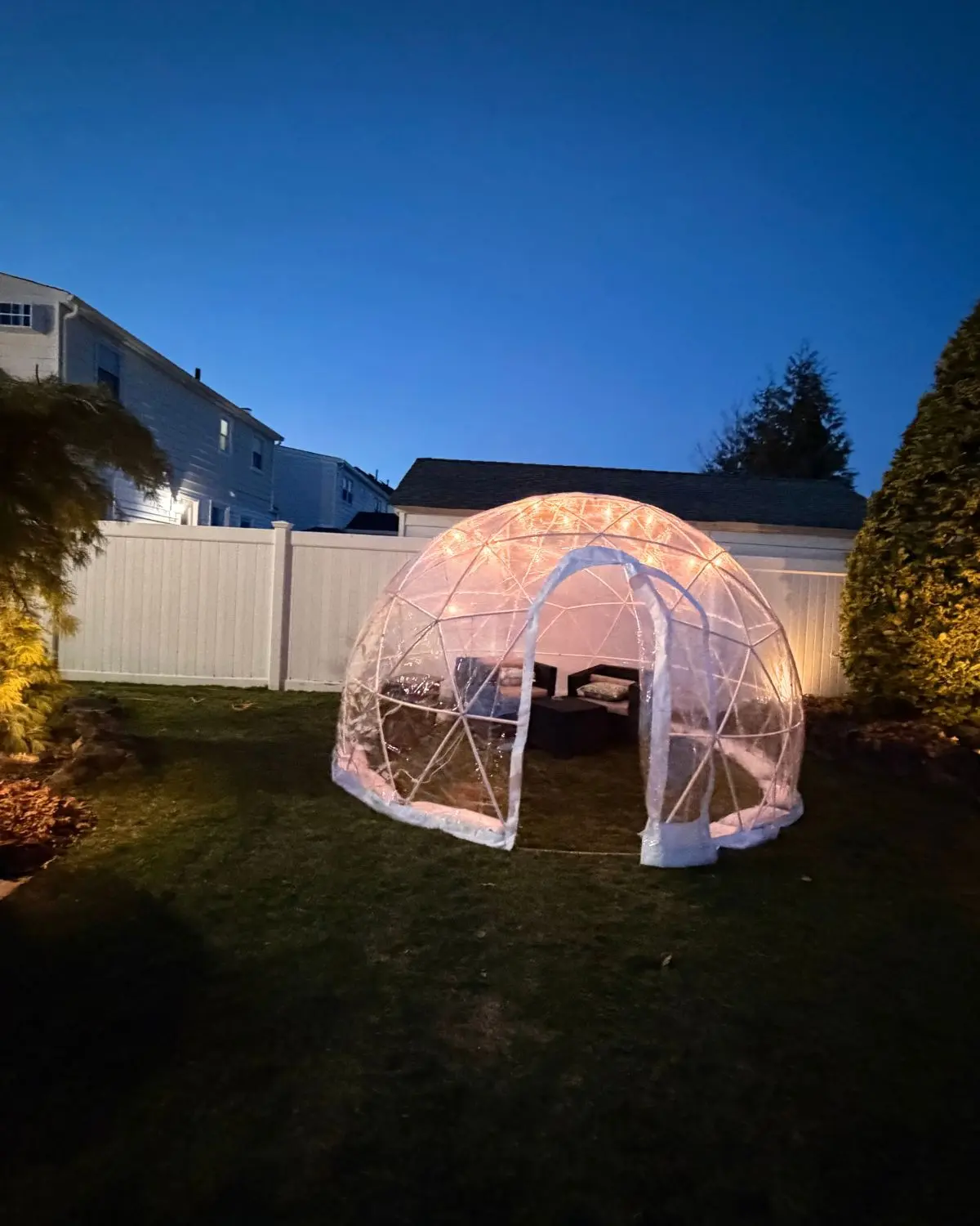Transparent geodesic dome igloo illuminated with string lights, set up on a backyard lawn at dusk with outdoor furniture inside, surrounded by white fence and trees.