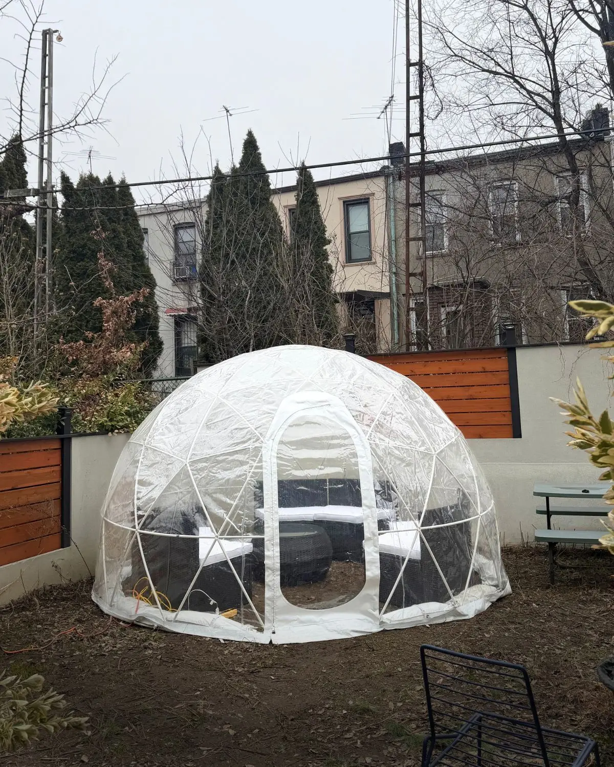 Transparent geodesic dome igloo set up in a backyard with patio furniture inside, surrounded by trees, plants, and residential buildings in the background.