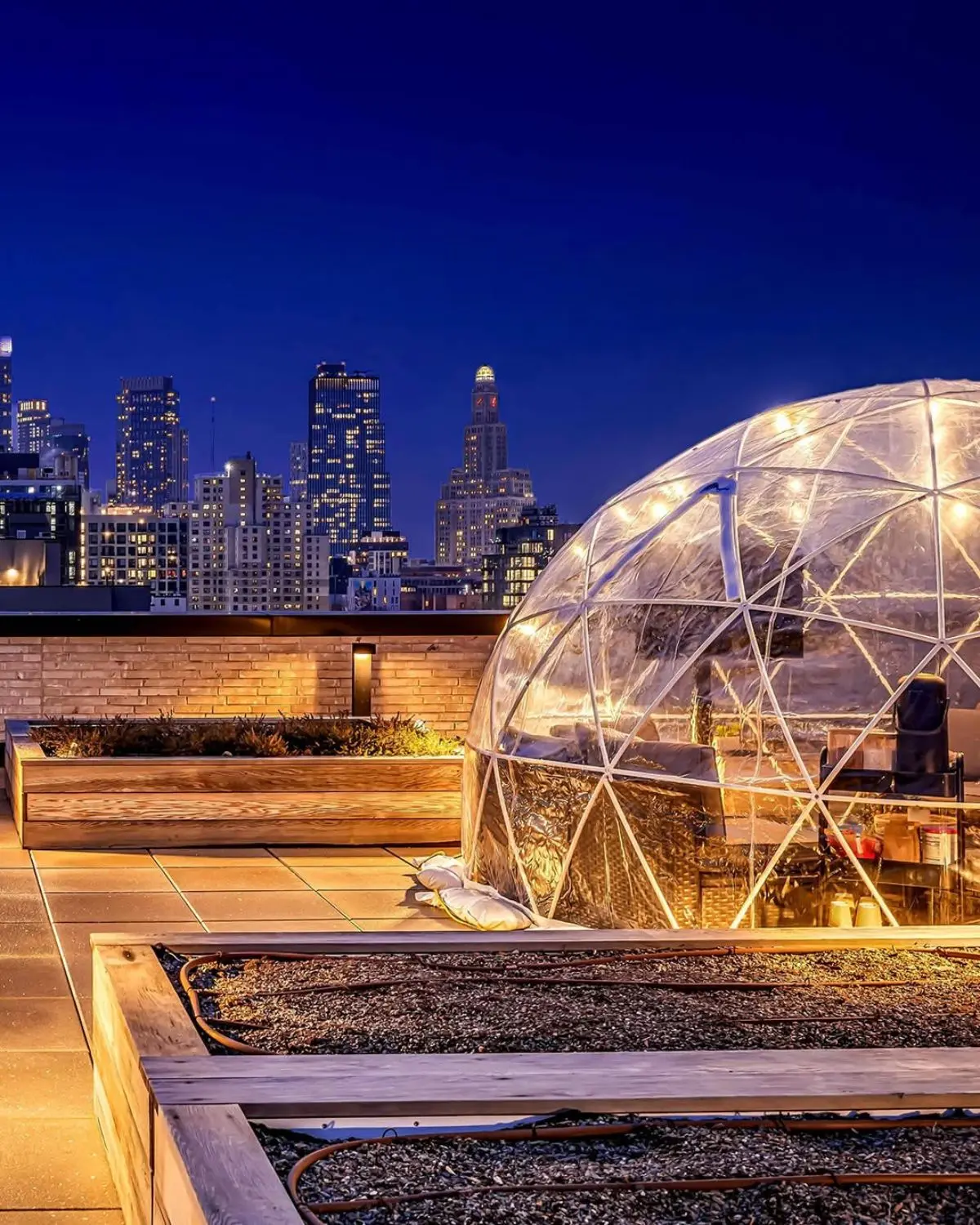 Illuminated transparent geodesic dome on a rooftop patio at night with city skyline in background, wooden planters, and tiled floor.
