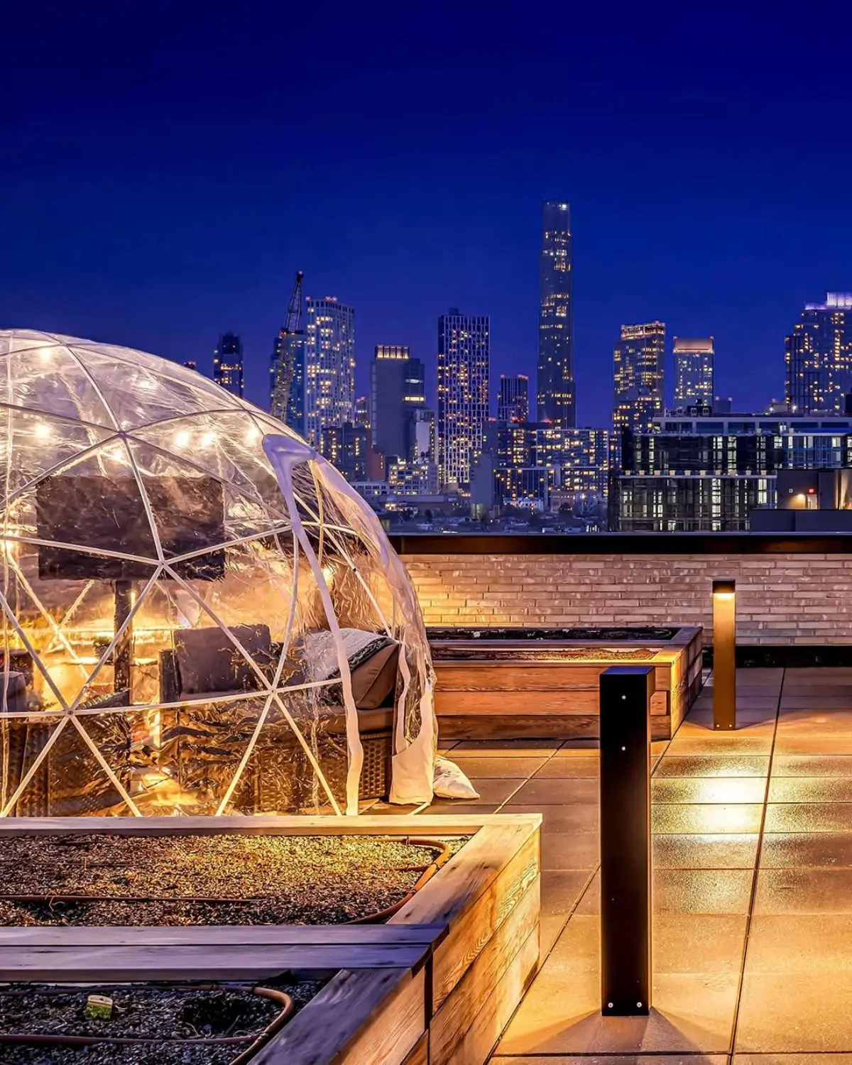 Transparent geodesic dome igloo lights on rooftop at night with city skyline in the background, featuring cozy seating inside and warm ambient lighting.