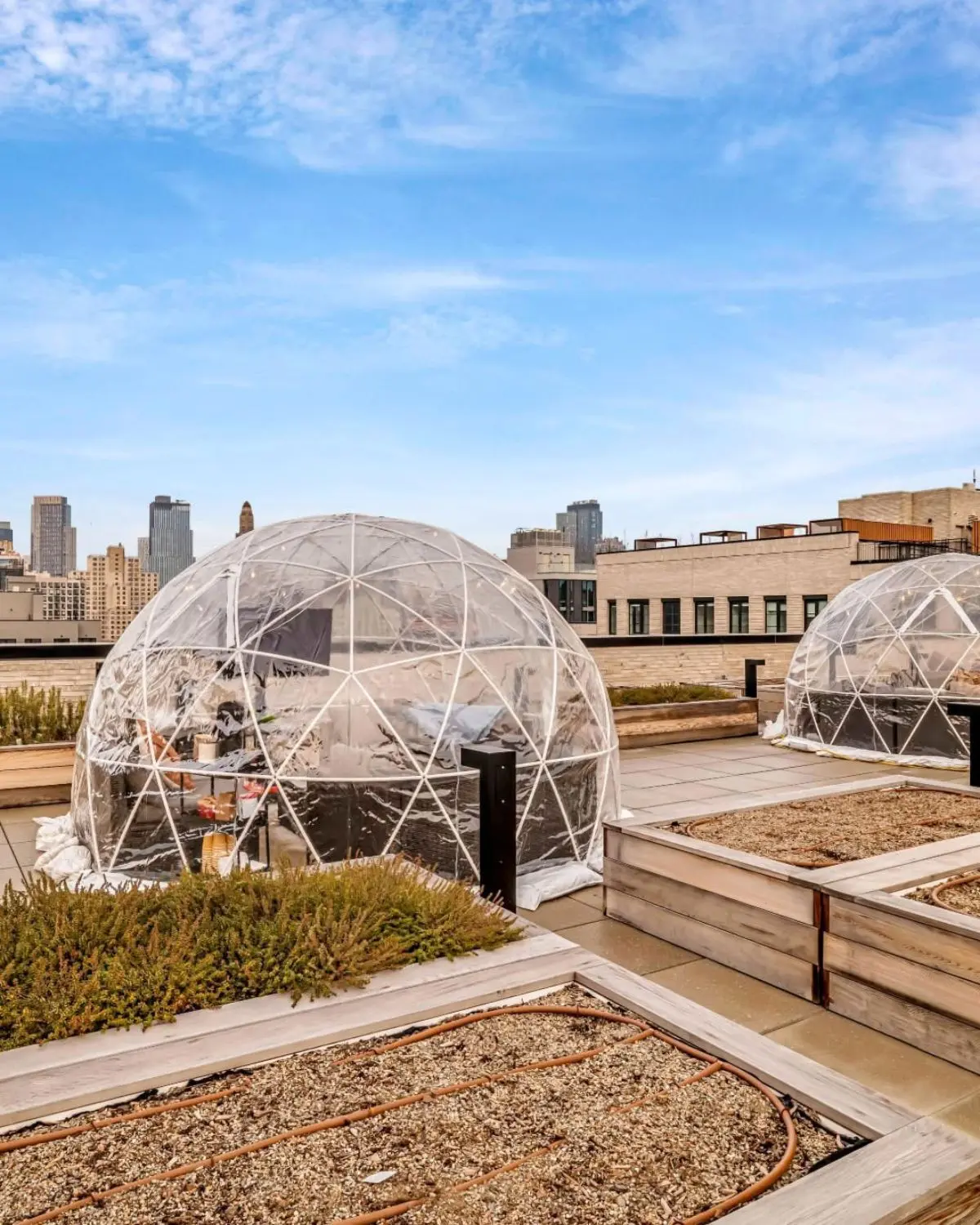Two transparent geodesic dome igloos on a rooftop terrace with seating inside, surrounded by raised garden beds and city buildings in the background under a partly cloudy blue sky.