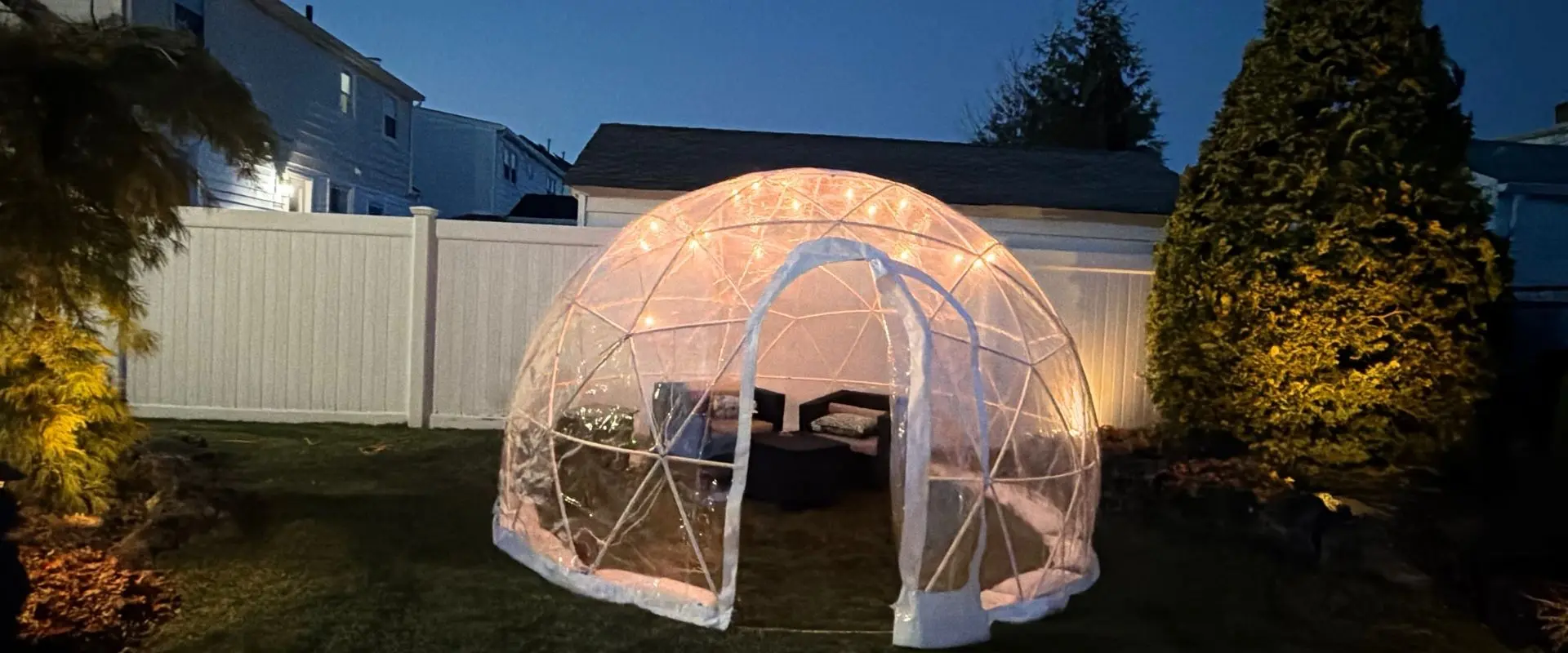 Heated clear igloo rental with lounge furniture and string lights set up in a Long Island backyard at night.